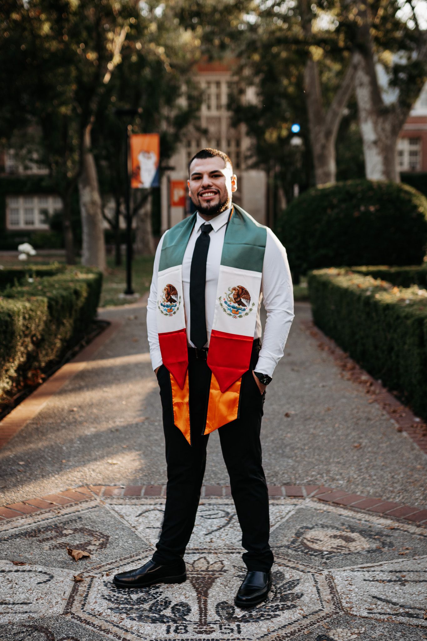 Mexican flag draped over shoulders walking on decorative stone pathway with trees in autumn scene.