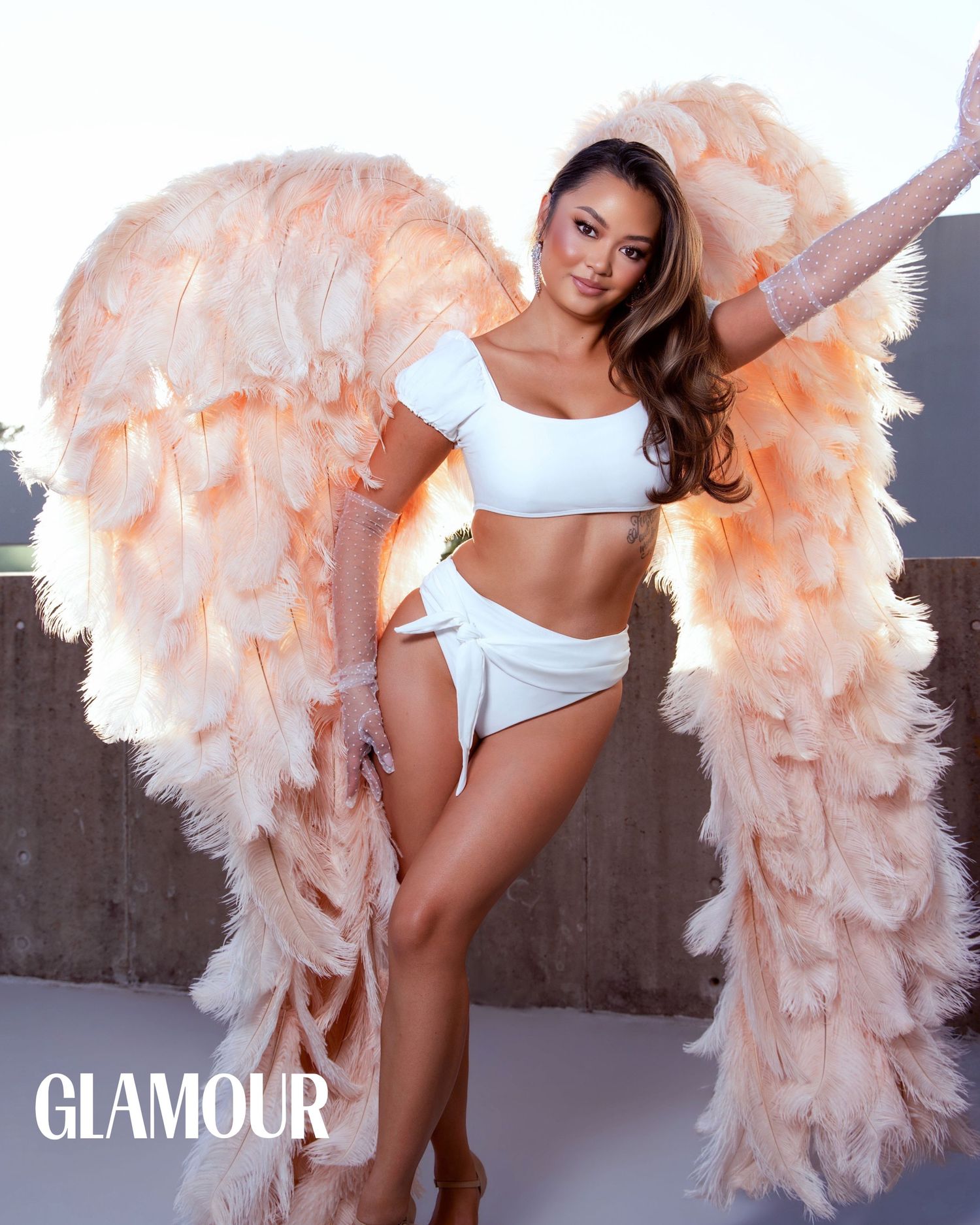 Model poses in white bikini with large pink feathered angel wings for Glamour magazine shoot.