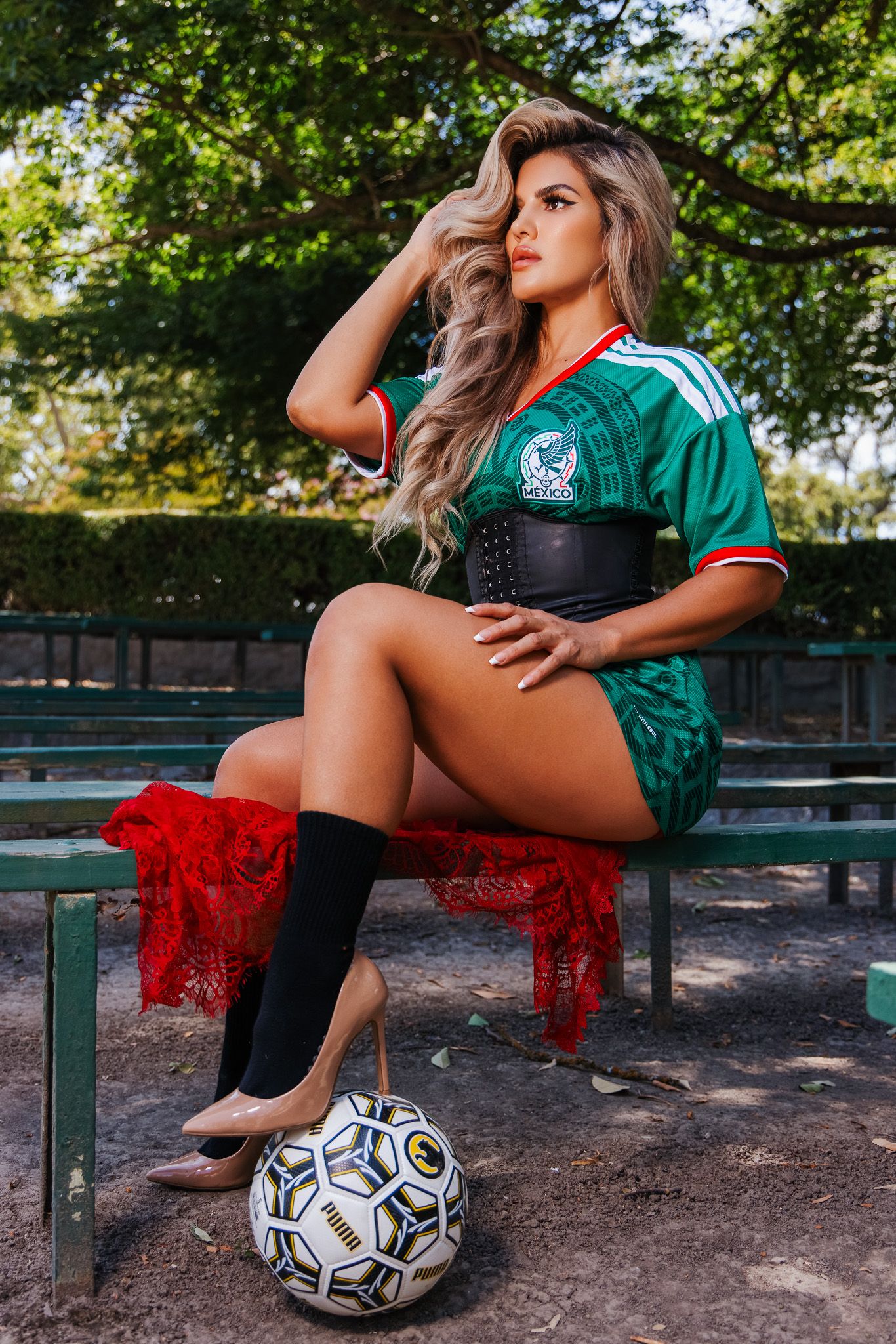 Woman in Mexico soccer jersey and black socks sits on bench with soccer ball, wearing a black corset belt outdoors.