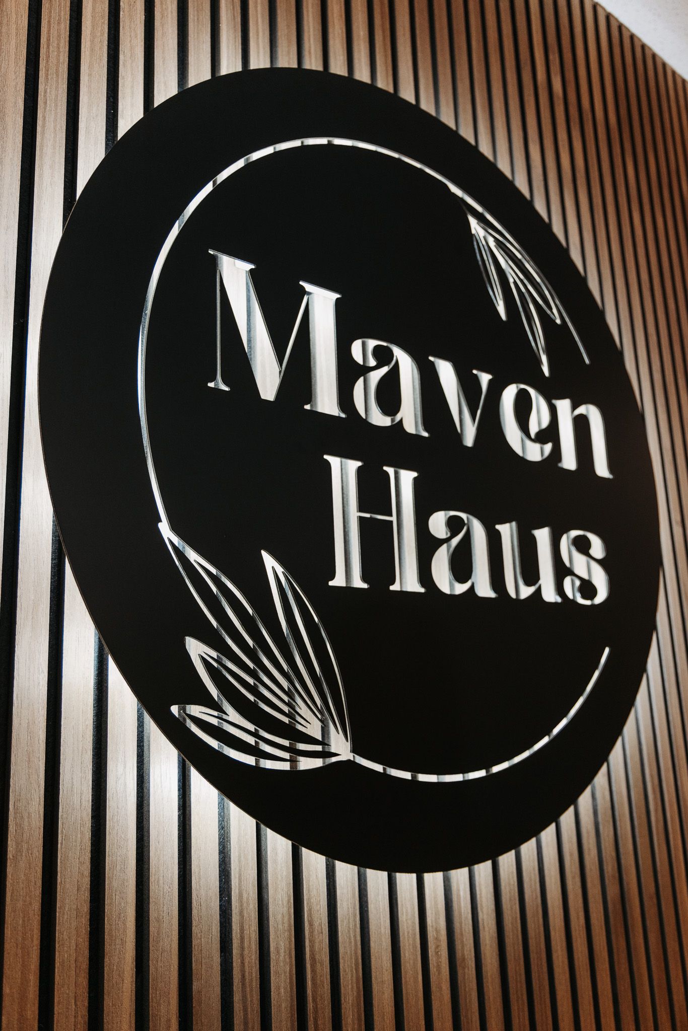 Elegant black oval Maven Haus logo sign mounted on a wood slat wall with botanical leaf accents.