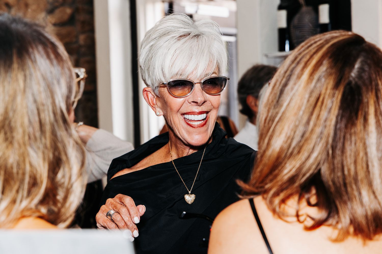 Older woman with short gray hair and sunglasses laughing joyfully at a social gathering wearing a black outfit.