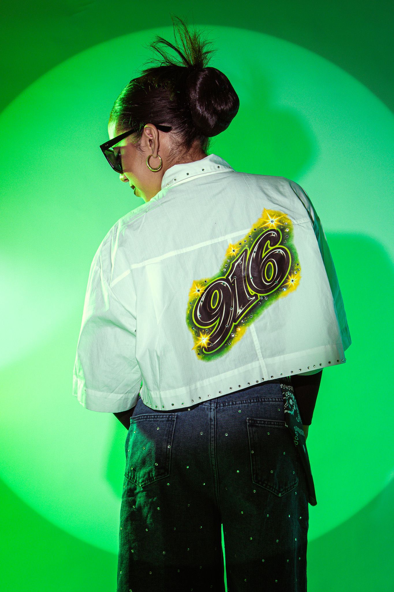 Person wearing white cropped jacket with '916' graphic print and studded jeans, posing against green background.