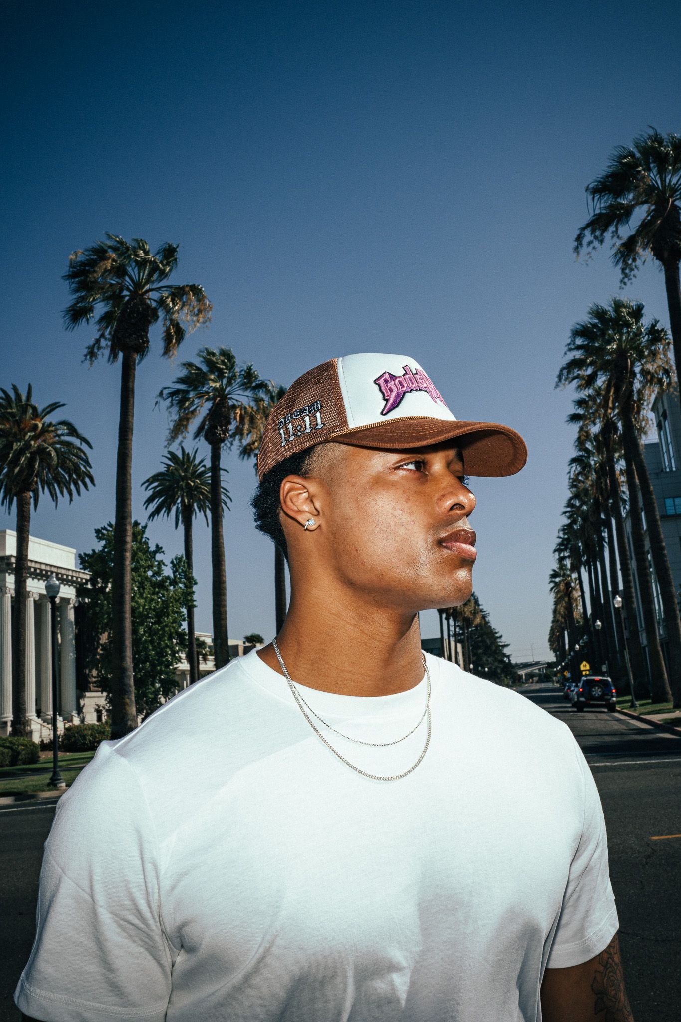 Young Black man in white tee and brown snapback cap poses on palm tree-lined boulevard under clear blue sky.