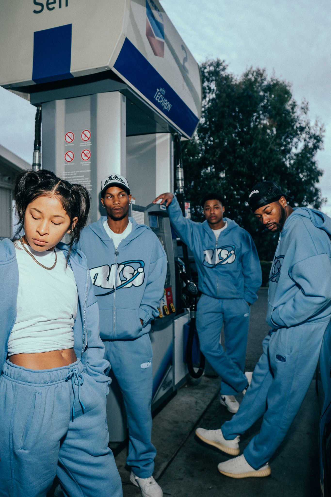 Four people wearing matching blue hoodies and sweatpants posing at a gas station in a stylish streetwear photoshoot.
