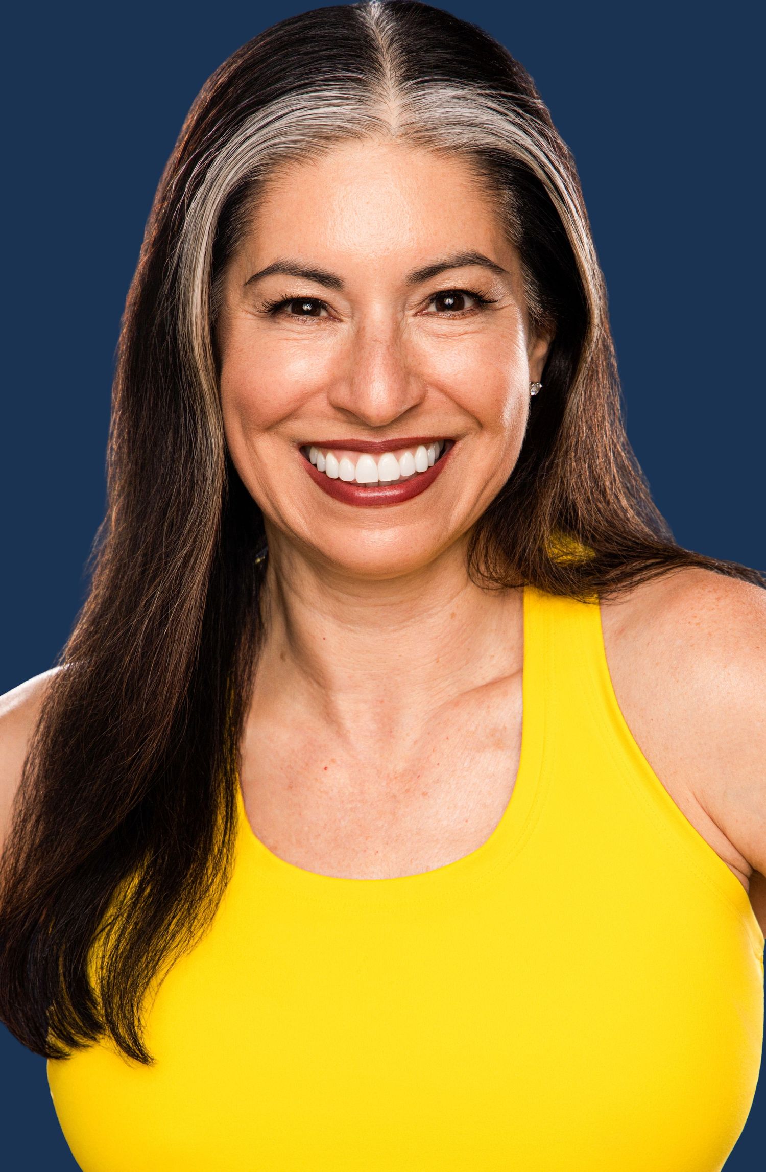 Smiling woman with long brown hair and gray highlights wearing a yellow tank top against a blue background.