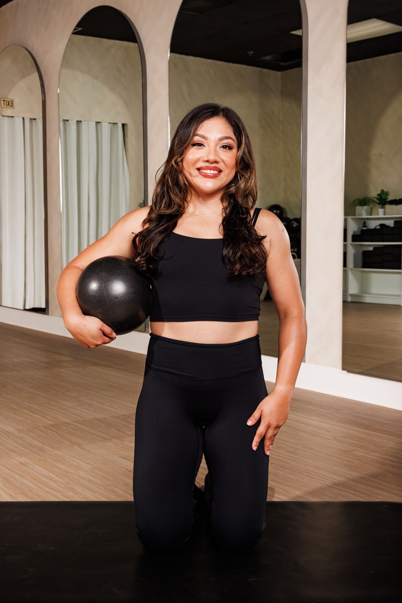 Smiling woman in black crop top and leggings holding a medicine ball in a gym.