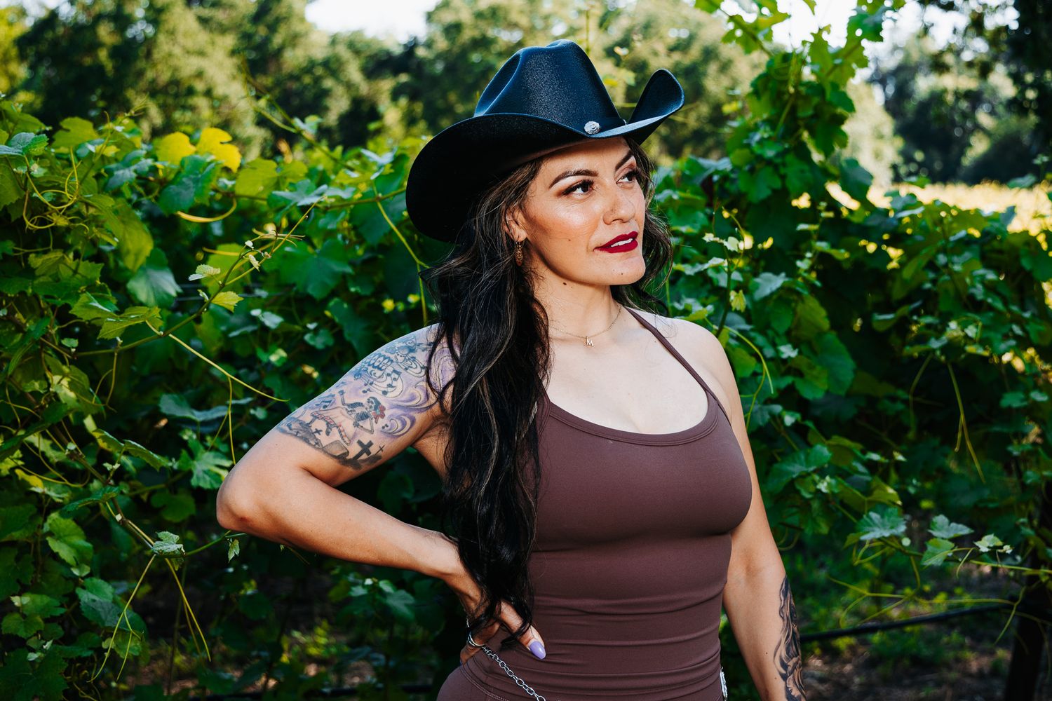 Woman in black cowboy hat with arm tattoo poses confidently among lush green vineyard leaves outdoors.
