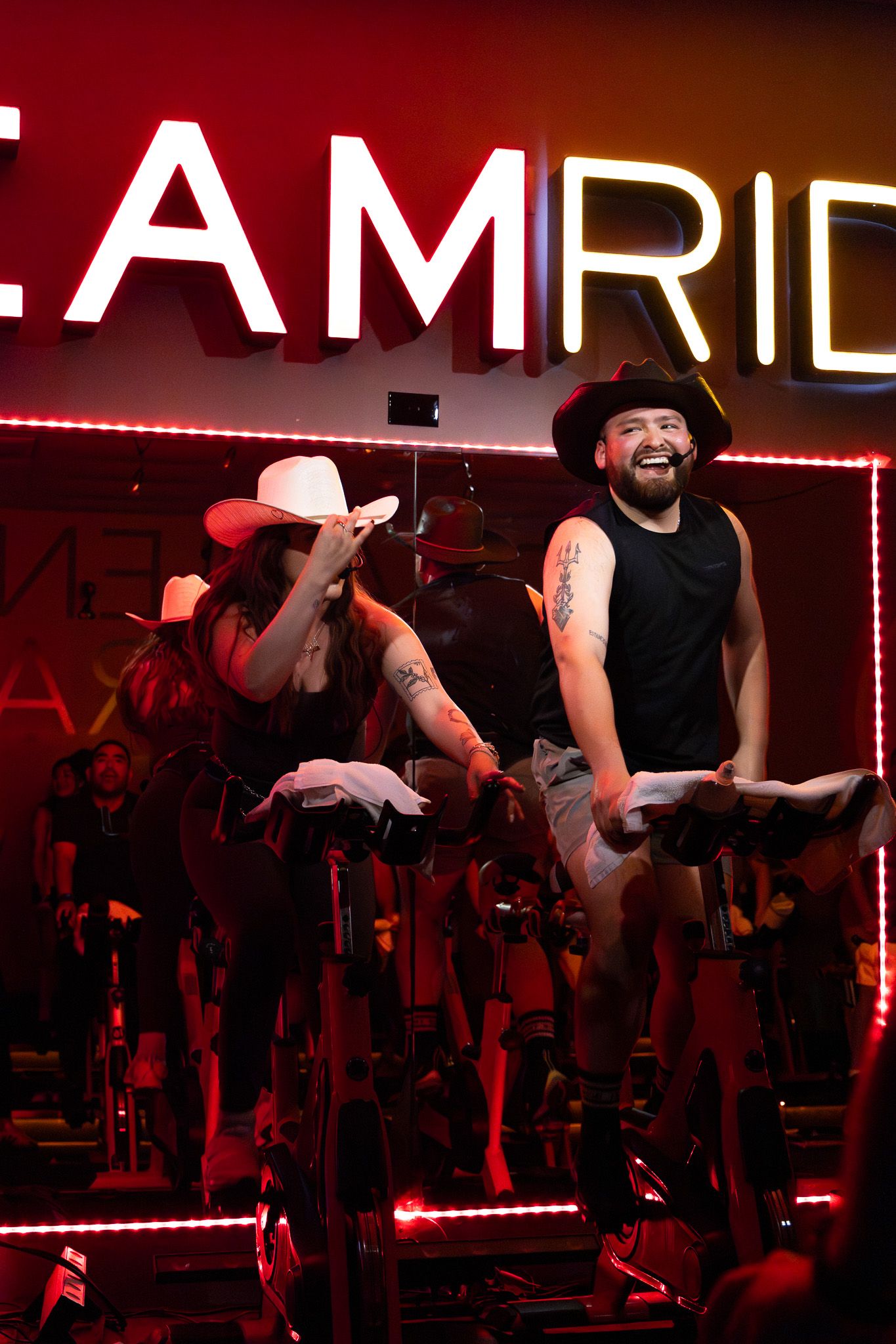 Two performers on exercise bikes engage in an energetic indoor cycling class under red lighting and AMRID signage.