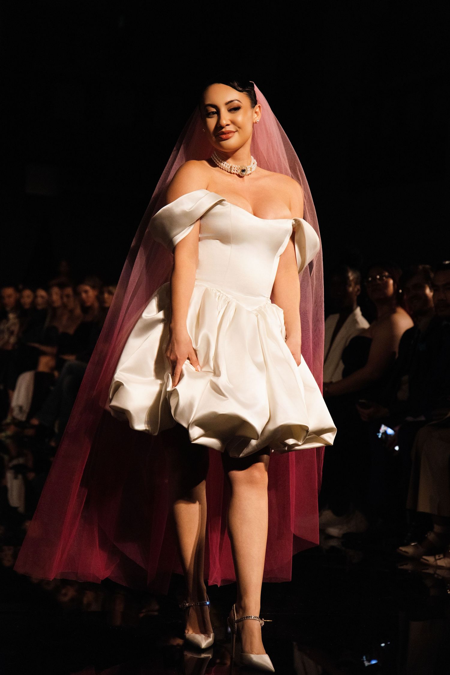 A model walks the runway in a short white dress with a flowing pink veil during a fashion show.