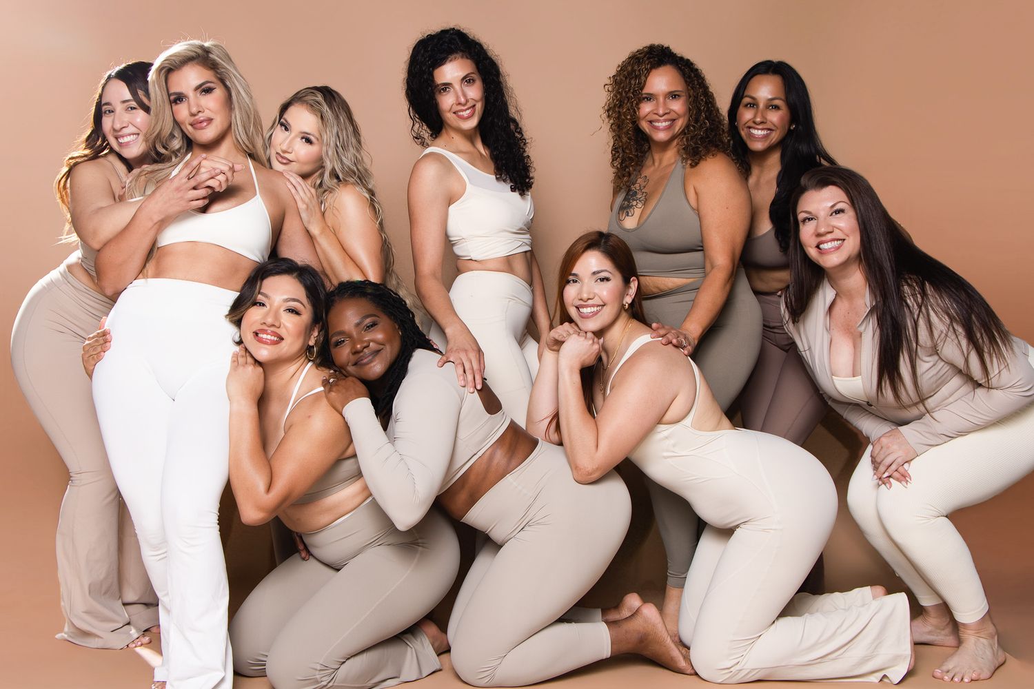 Diverse group of ten women in neutral activewear posing together on beige background.