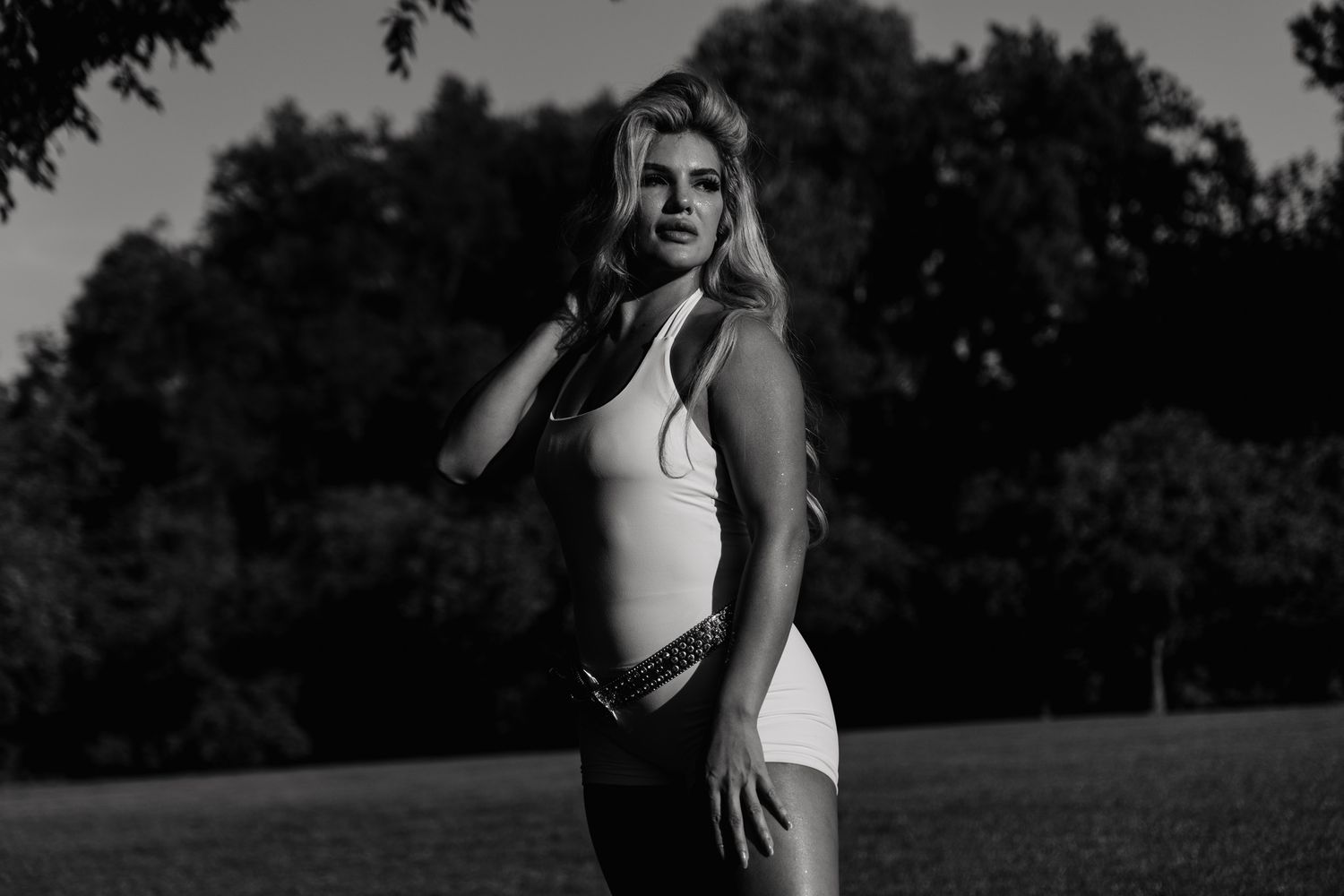 Black and white photo of a young blonde woman posing outdoors in a park wearing a white top and belt.
