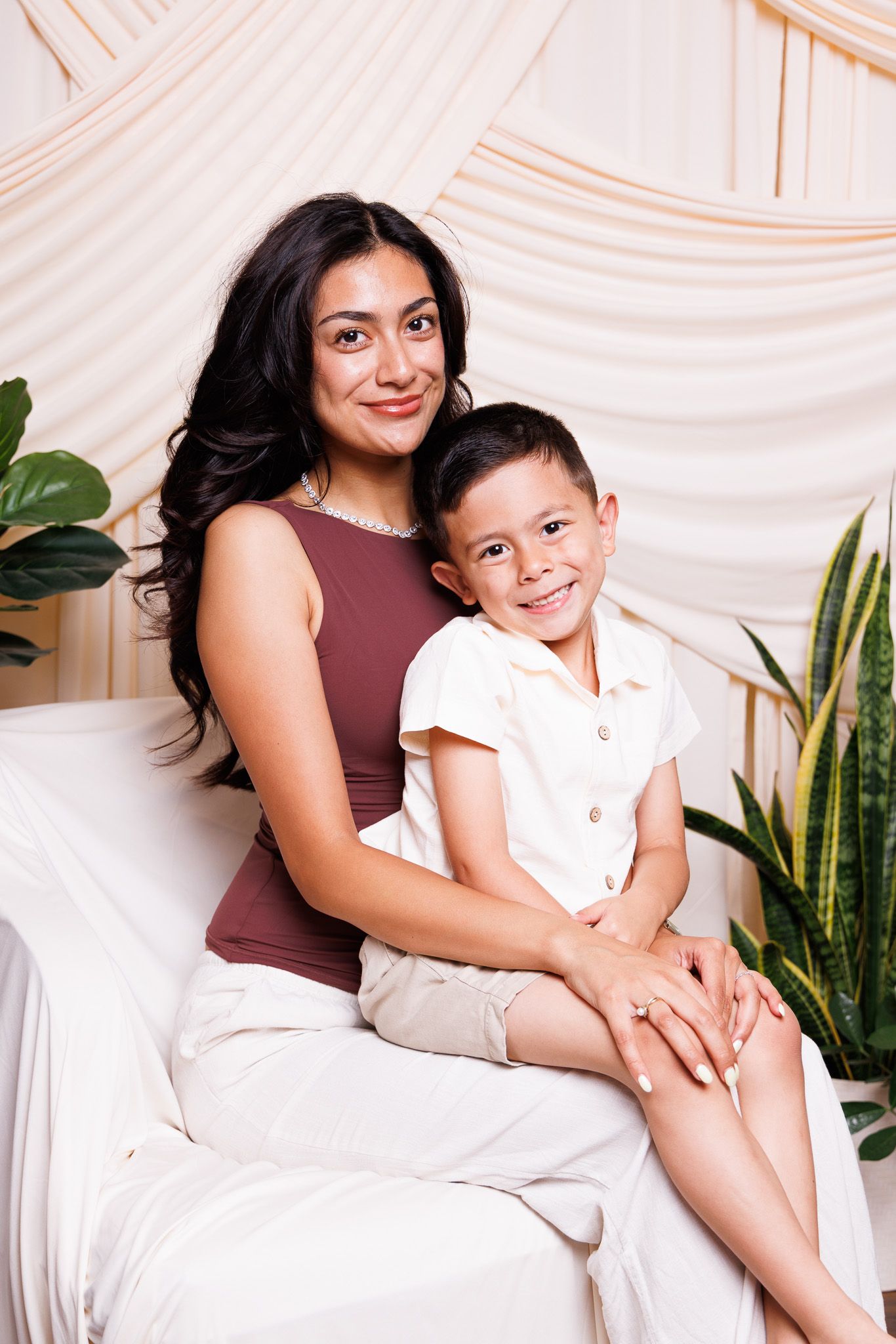 A woman in a brown top and white skirt sits smiling with a young boy in a white outfit on a couch.