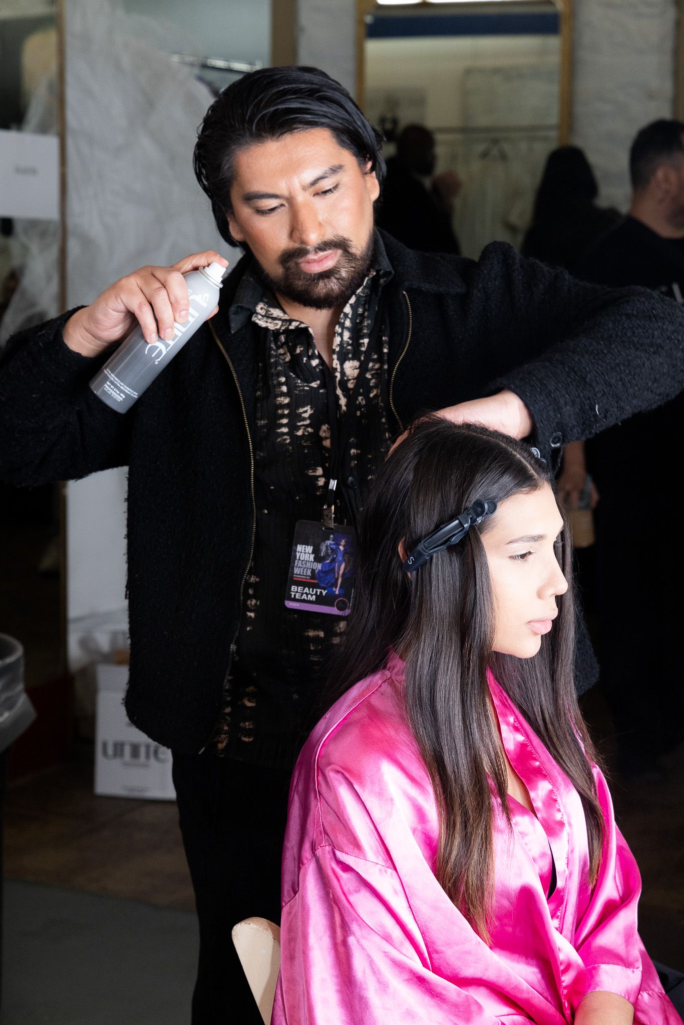 Stylist works on model's hair backstage while model wears pink silk robe.