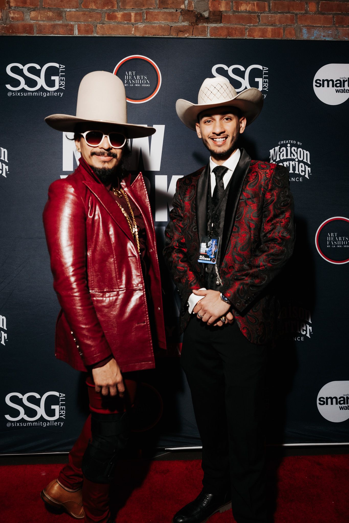 Two people wearing cowboy hats and stylish suits pose on a red carpet at a fashion event.