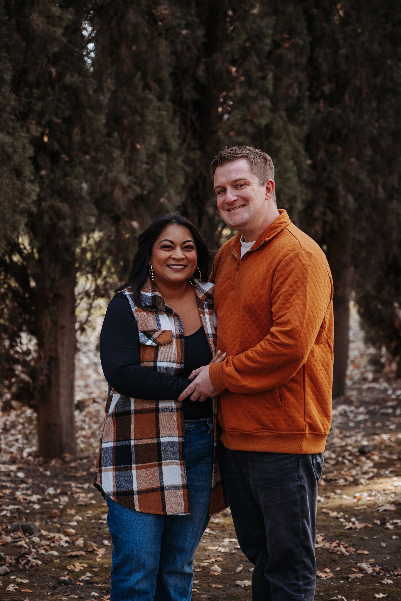 Couple in autumn attire share playful moments in a wooded area with fallen leaves.