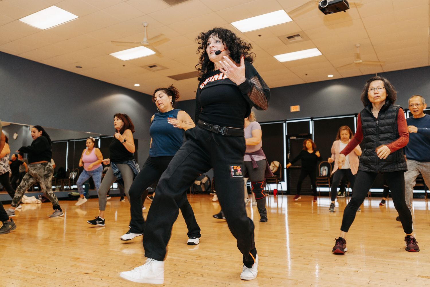 A group of people practicing dance moves in a studio with hardwood floors and ceiling lights.