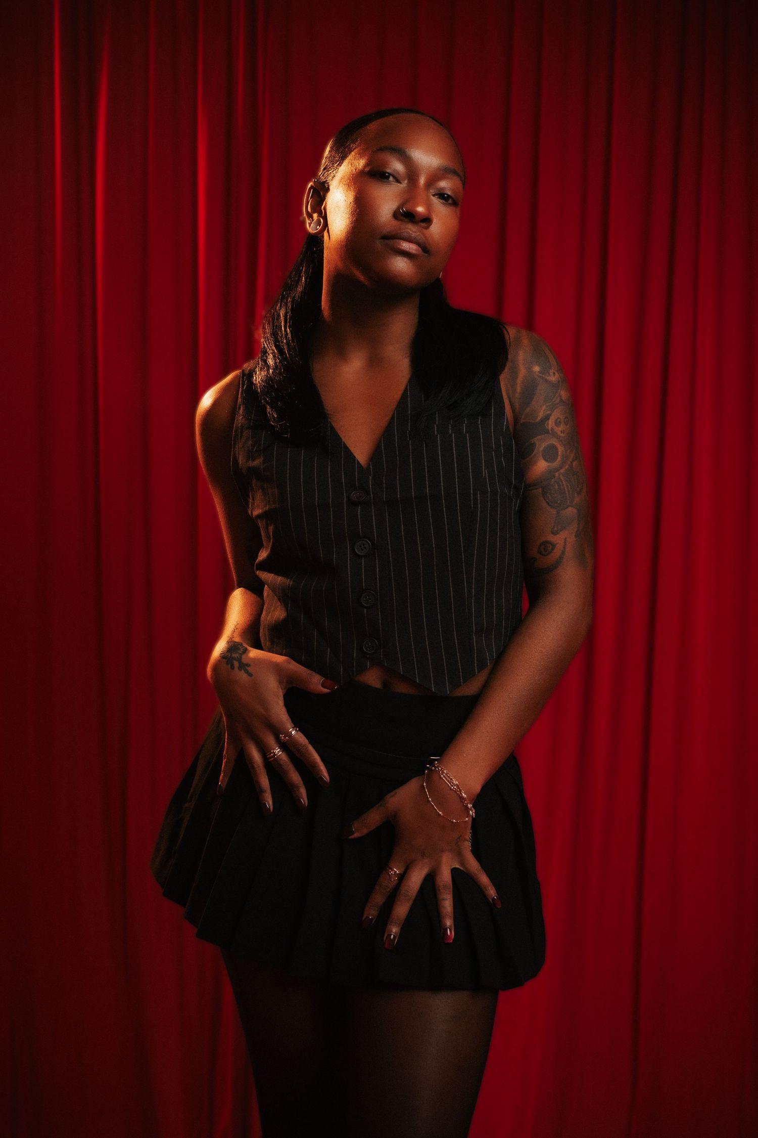 A person in a pinstripe vest and dark skirt poses dramatically against a red curtain backdrop with moody lighting.