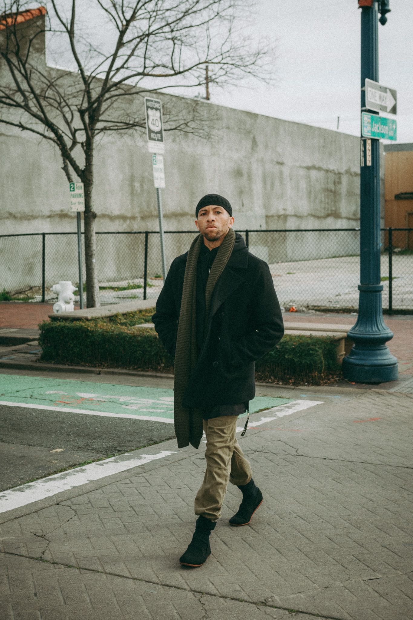 Person wearing black winter coat and khaki pants walking on urban sidewalk in winter setting.
