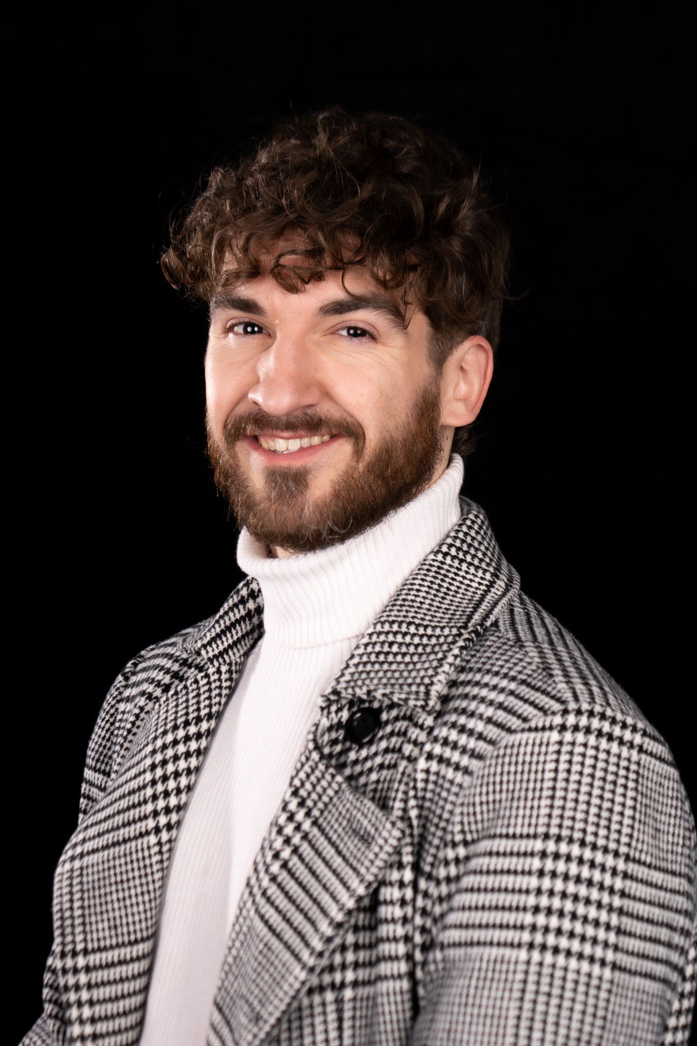 Professional studio portrait against black background wearing plaid blazer and white turtleneck sweater.