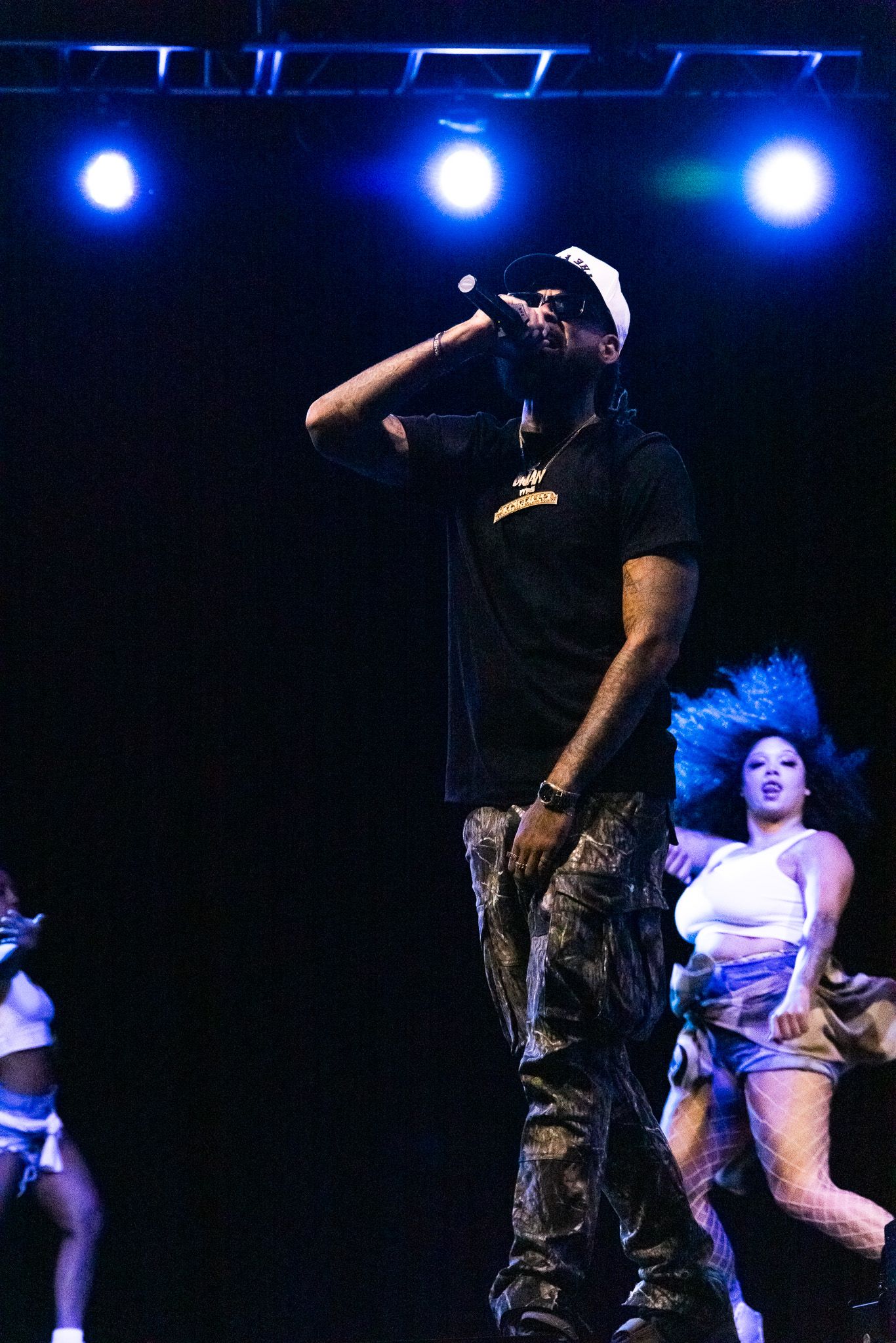 Performer in black shirt and camo pants singing on dark stage with blue spotlights and dancers in background.