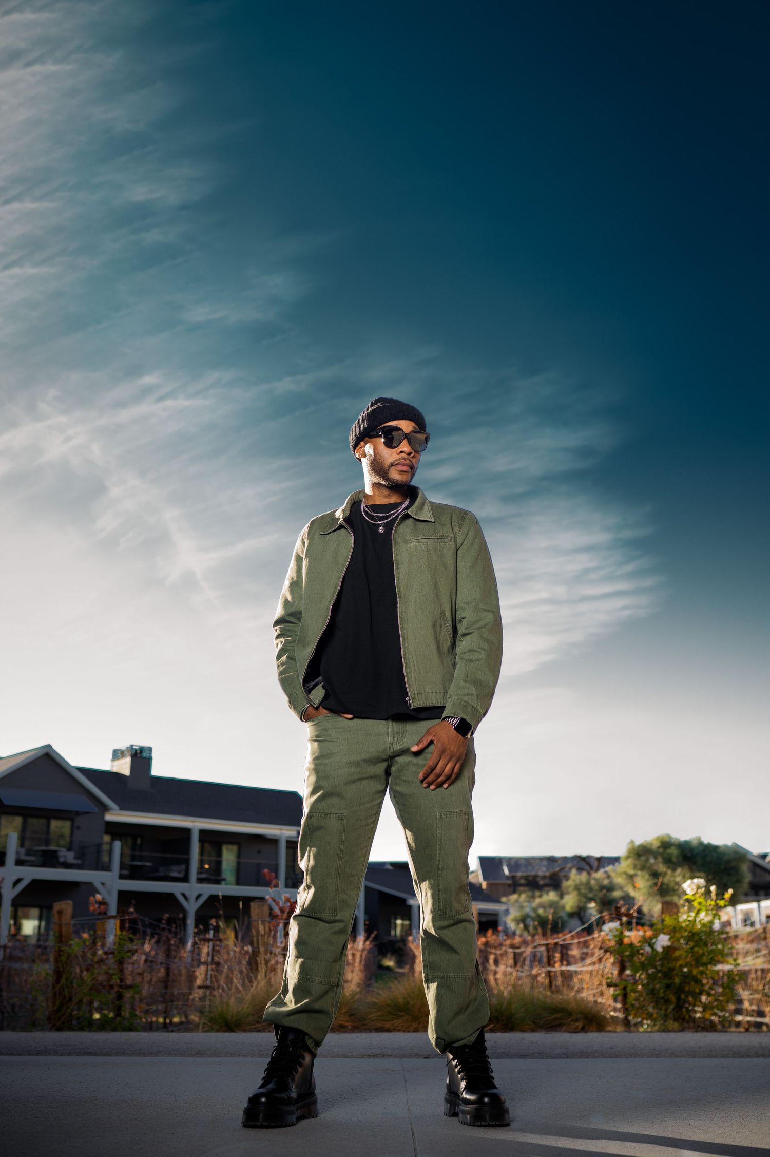 A stylish man in olive green jacket and cargo pants with sunglasses poses outdoors under a dramatic blue sky.