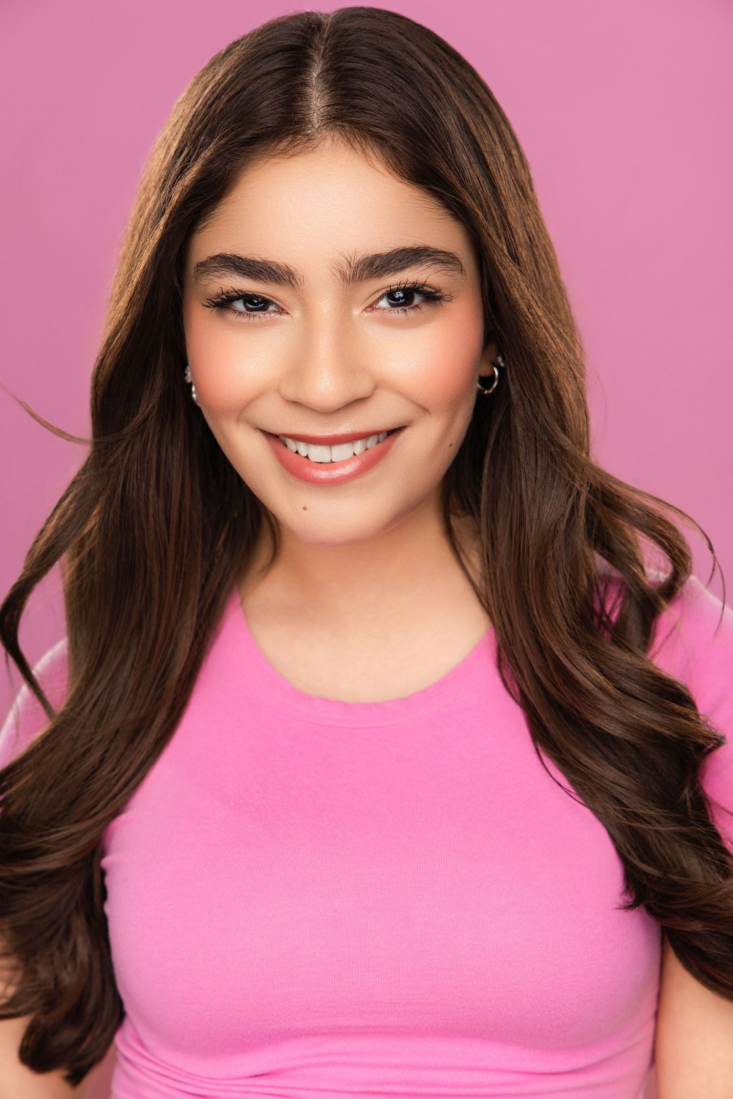 A smiling person with long dark hair wearing a bright pink top against a pink background.