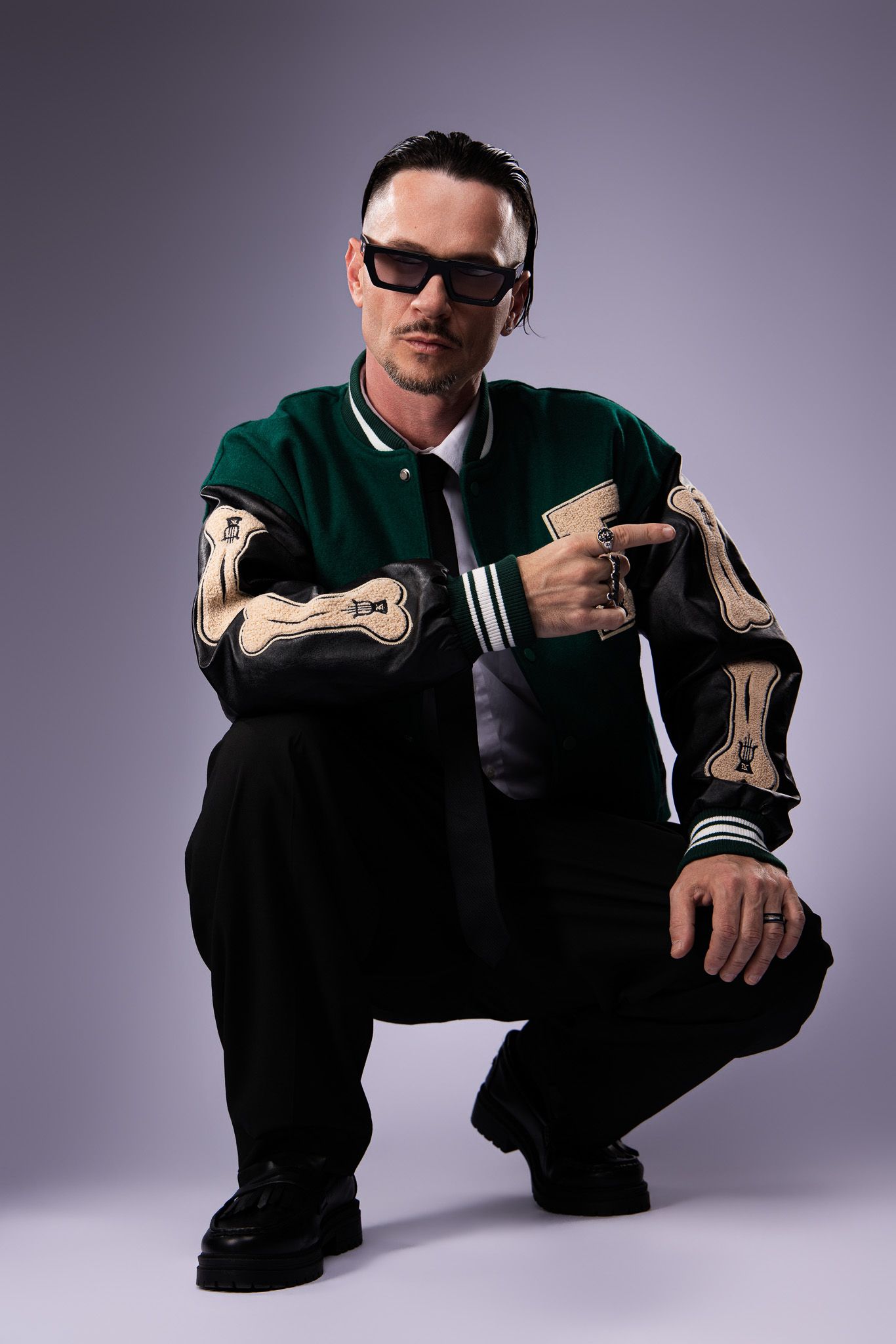 Person in sunglasses crouches wearing a green varsity jacket with bone designs on sleeves and black pants.