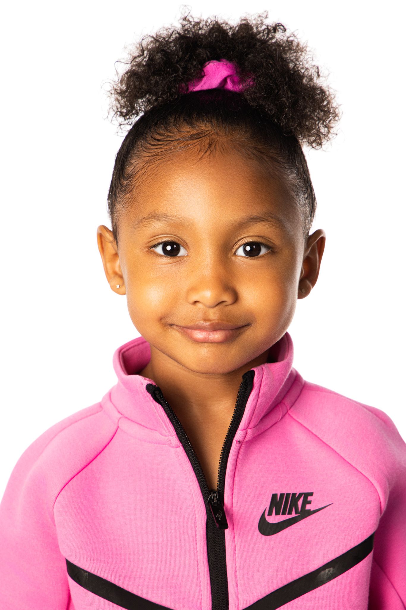 Young child wearing a pink Nike zip-up athletic jacket with curly hair styled in a bun with a pink hair accessory.