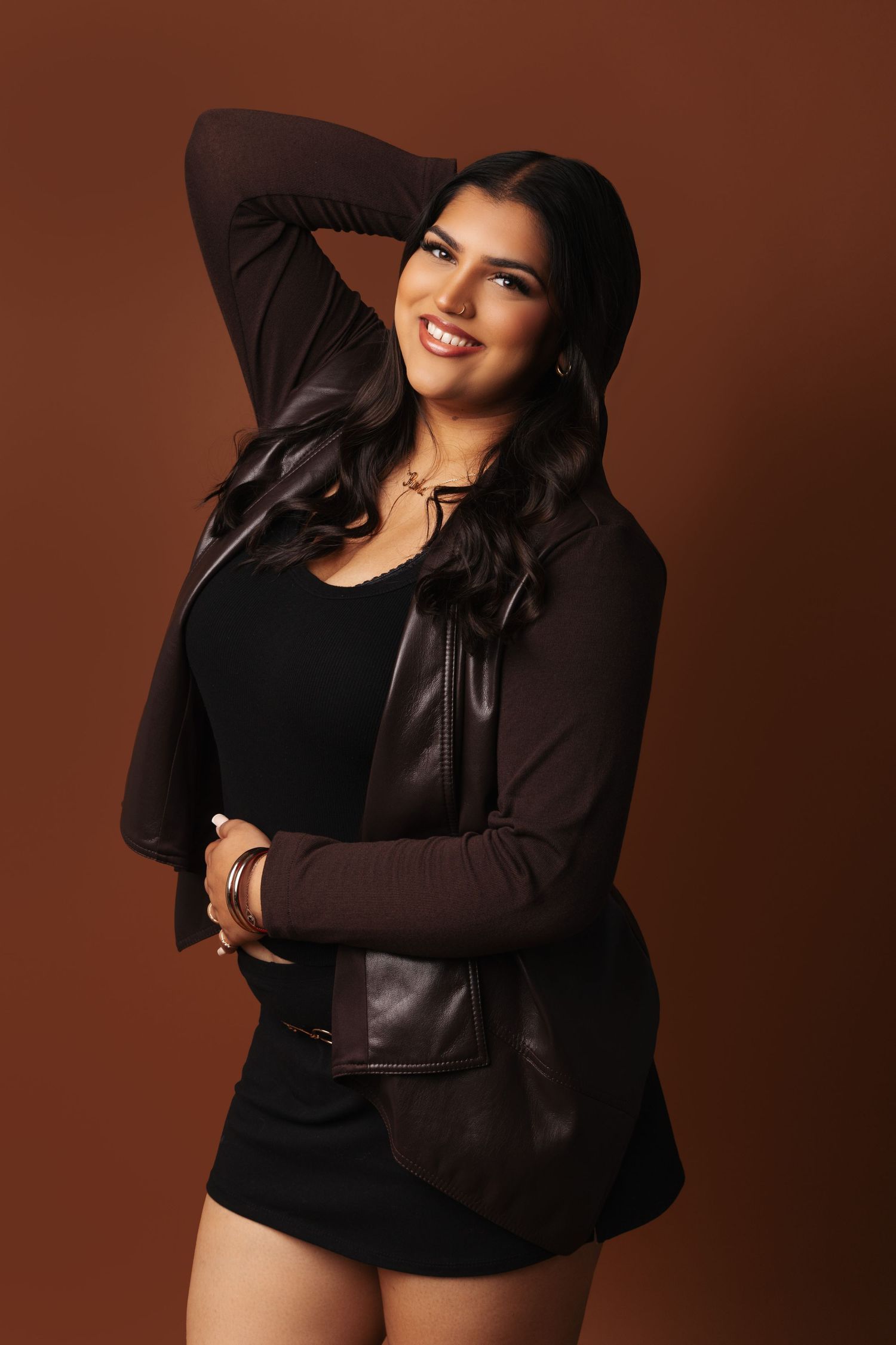Studio portrait against brown backdrop showing a woman in black outfit with leather jacket posing confidently.