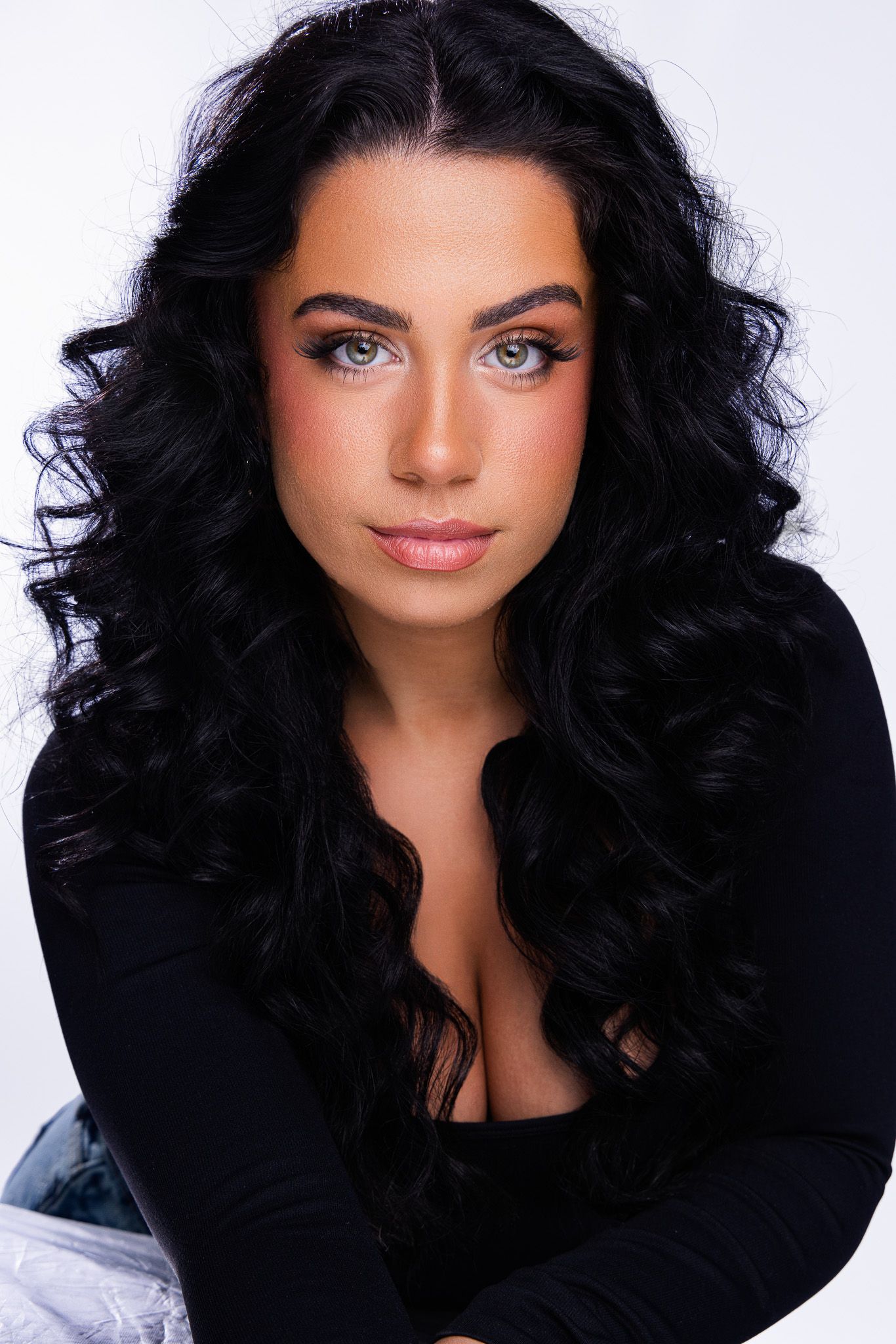 Young woman with long curly black hair and green eyes wearing a black top, posing against a white background.