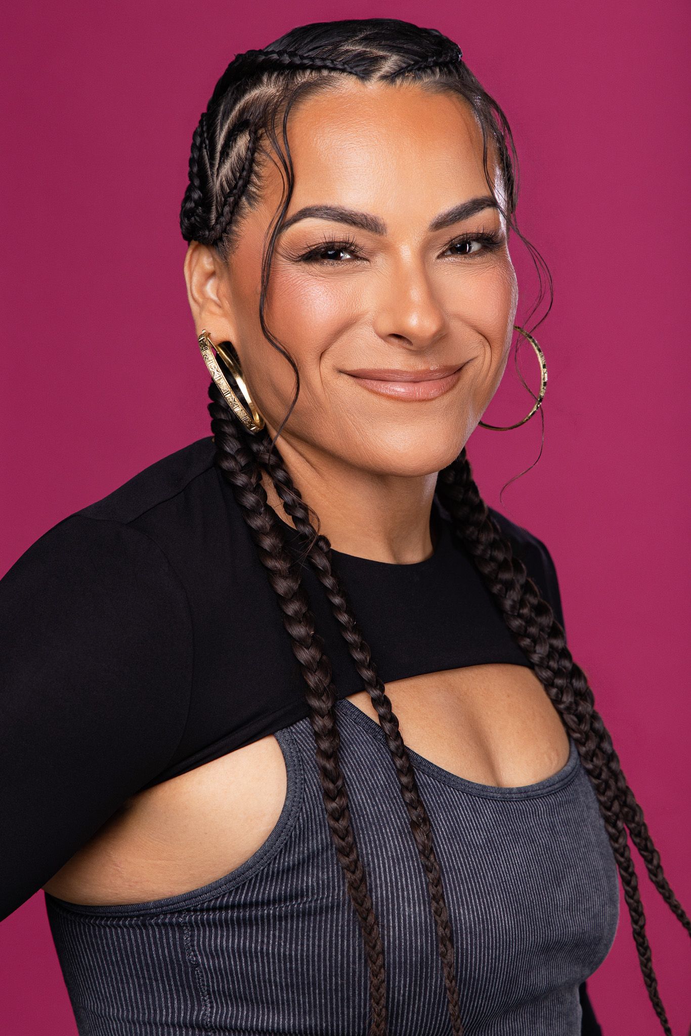Woman with braided hair and hoop earrings smiling against a pink background, wearing a gray top and black jacket.