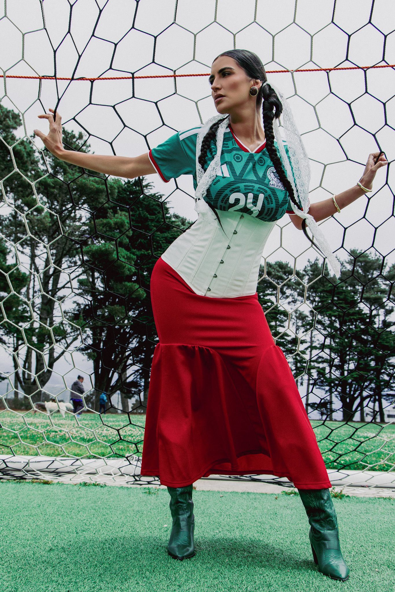 Woman in green jersey, white corset, red skirt, and green boots poses against a soccer goal net outdoors.