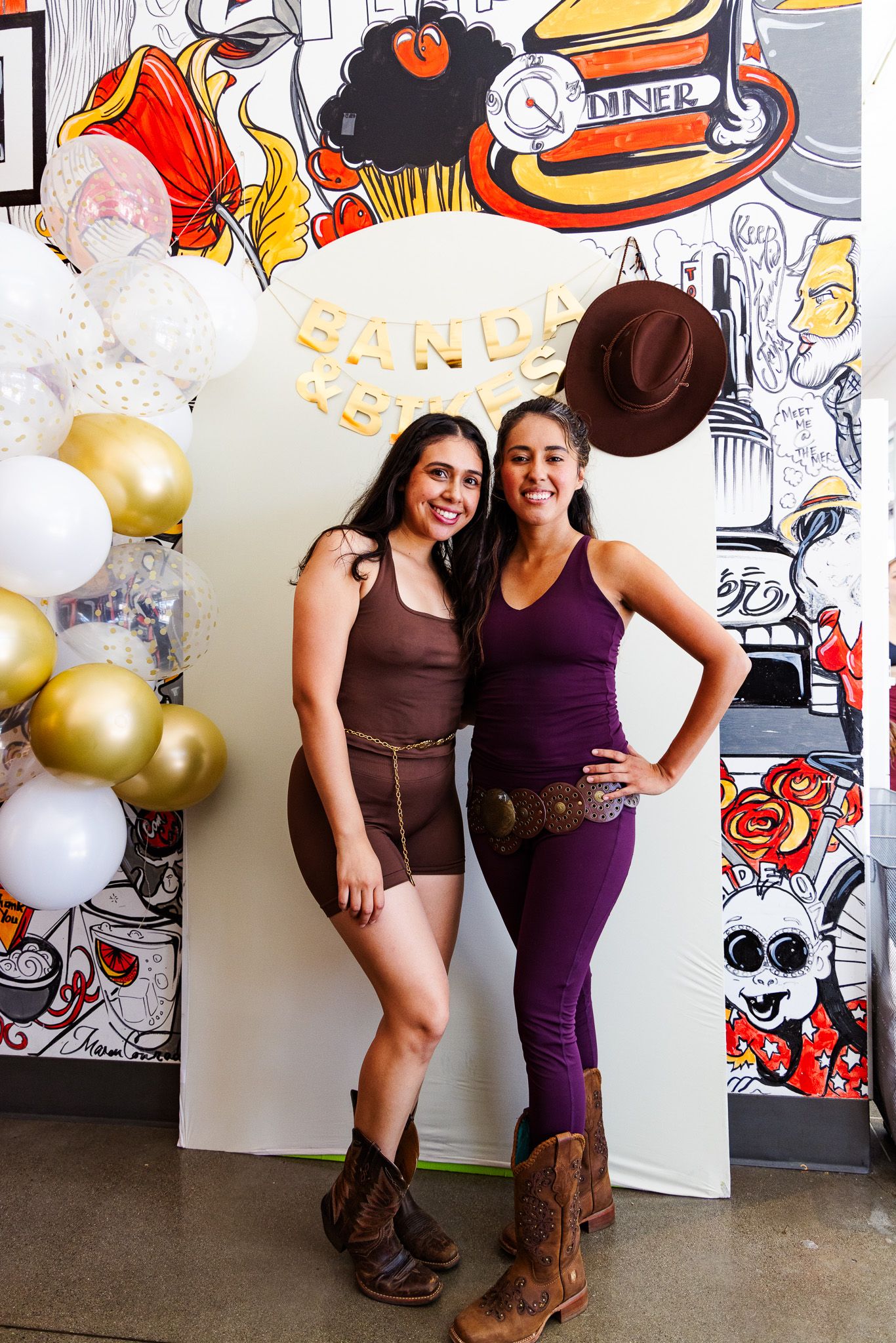 Two women in cowboy boots and western outfits pose at a festive party with balloons and colorful mural backdrop.