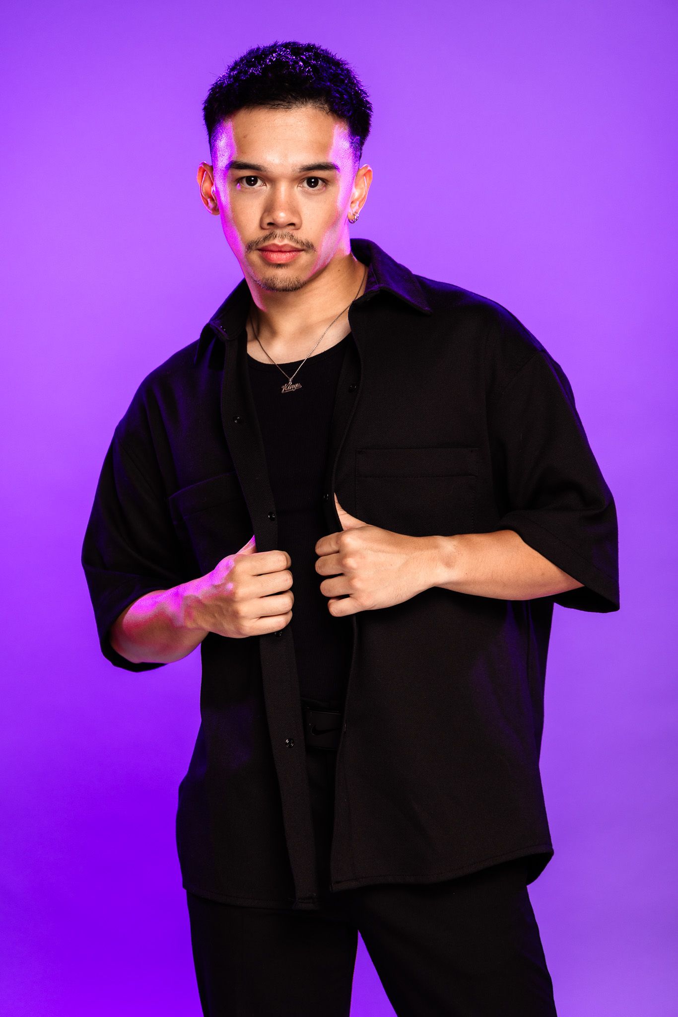 Young man in black outfit poses confidently against vibrant purple background, wearing necklace and earring.