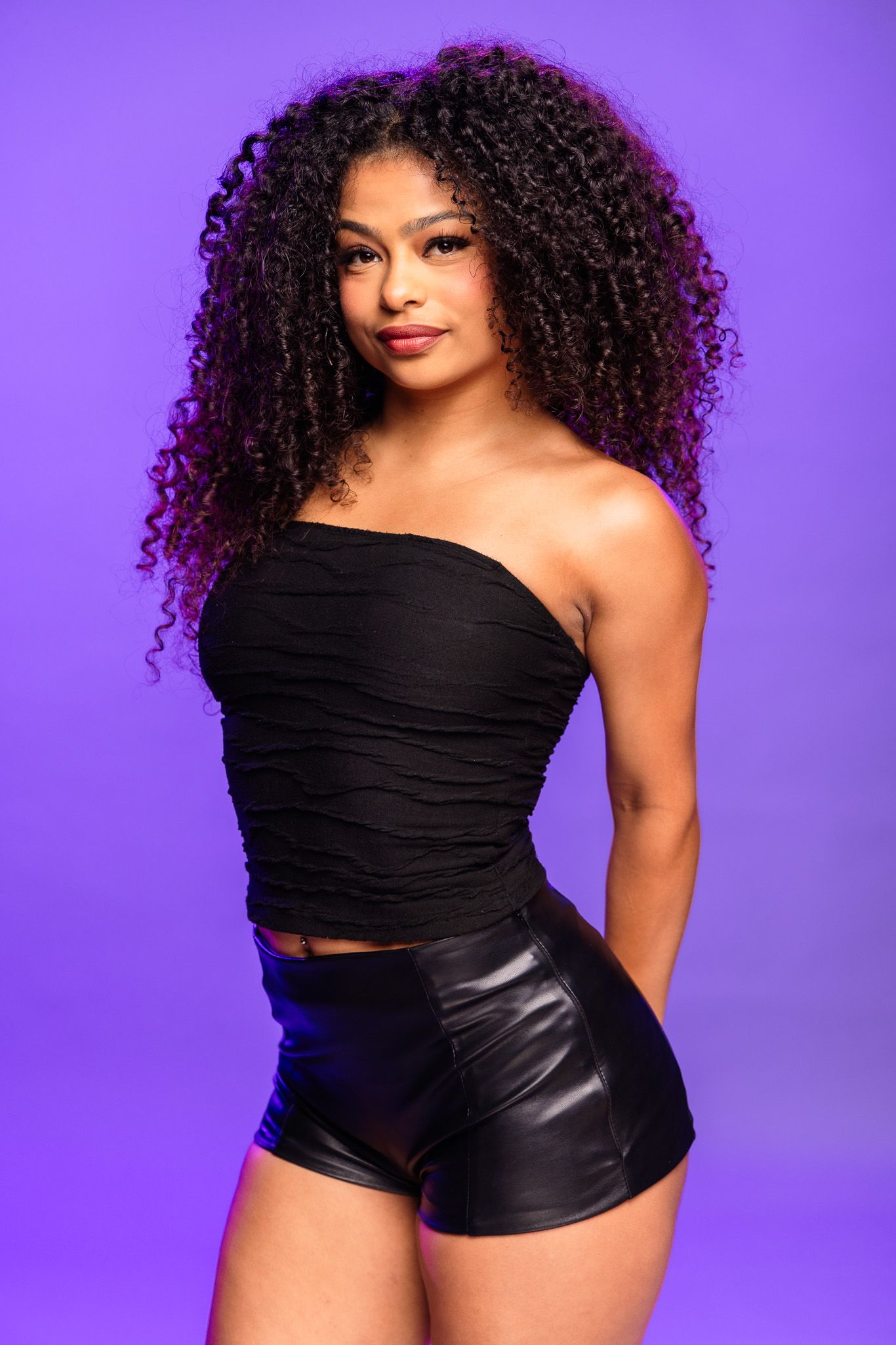 Young woman with voluminous curly hair wearing black tube top and leather shorts poses against purple background.