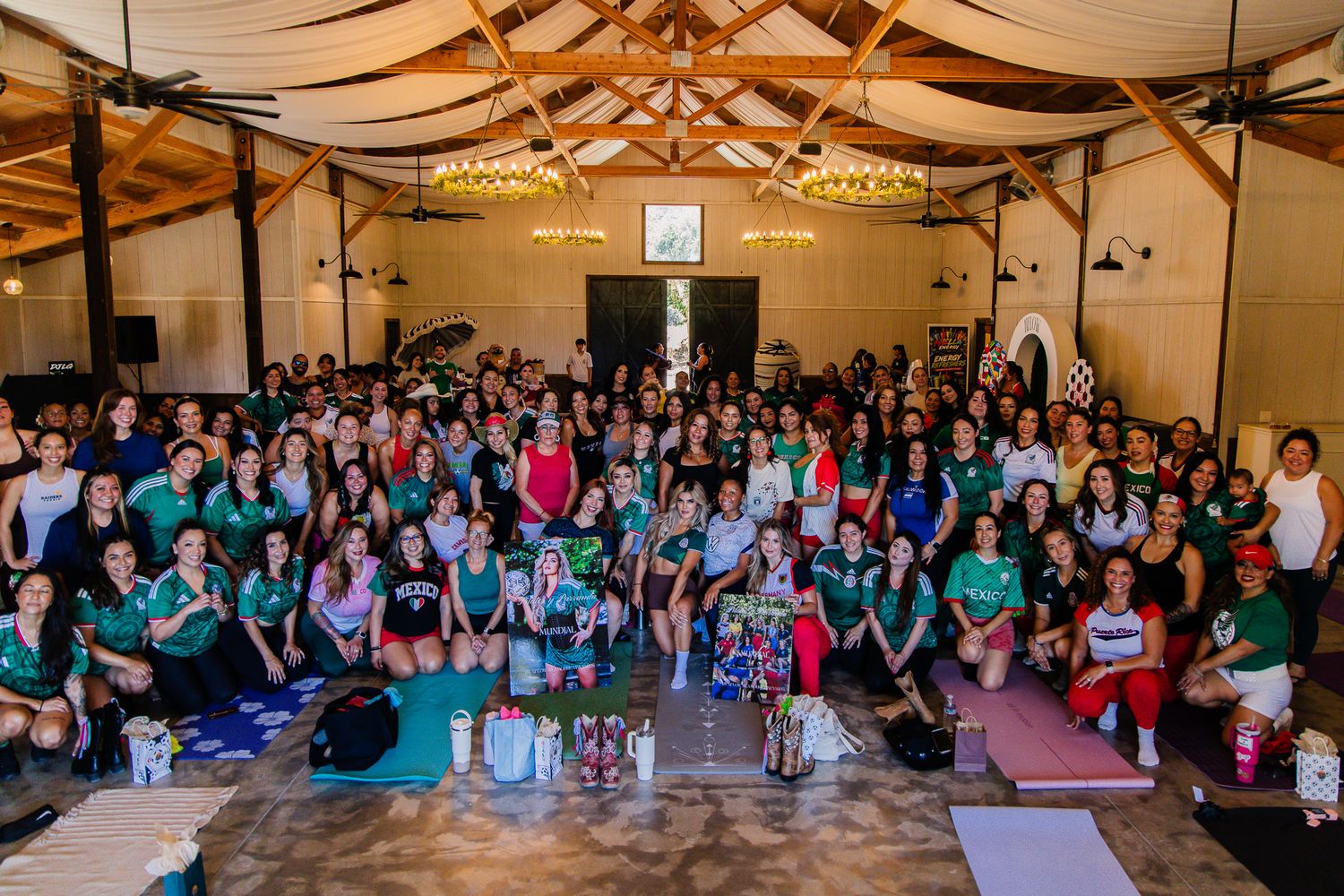 Large group of women in green shirts posing together inside a decorated wooden venue during a yoga or wellness event.