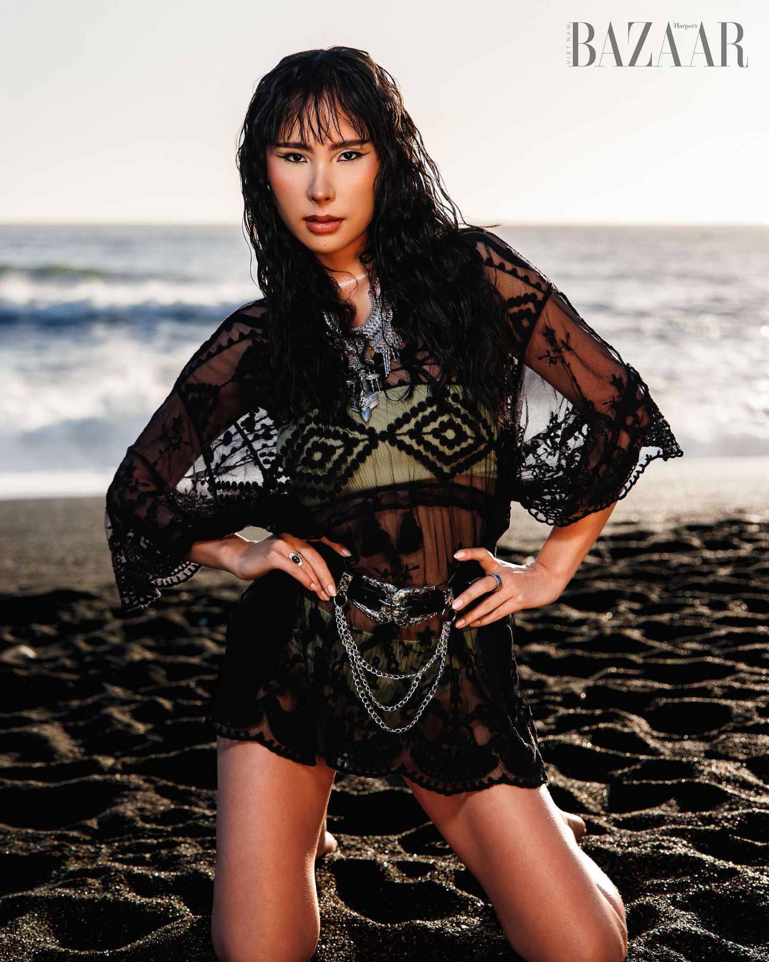 Woman in black lace cover-up with chain belt poses on sandy beach for Harper's Bazaar fashion shoot.