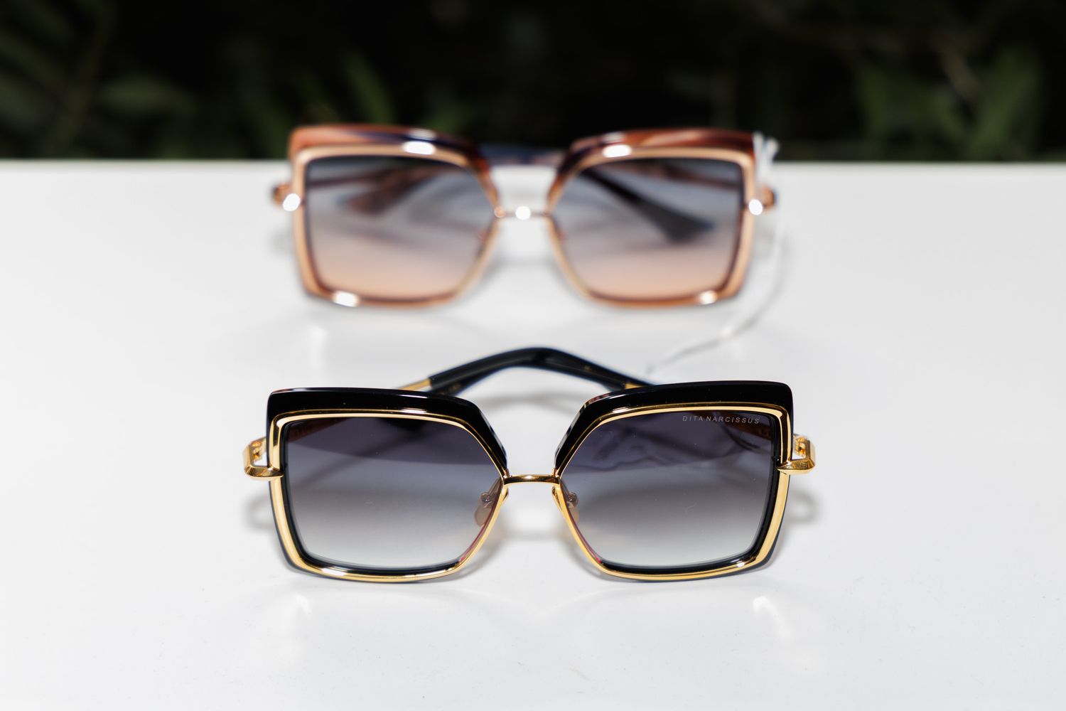 Two pairs of square luxury sunglasses with gold frames, one pink gradient and one black gradient, displayed on white surface.