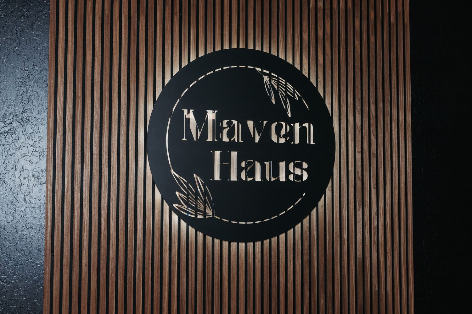 Black circular Maven Haus logo sign mounted on wooden slat wall panel.