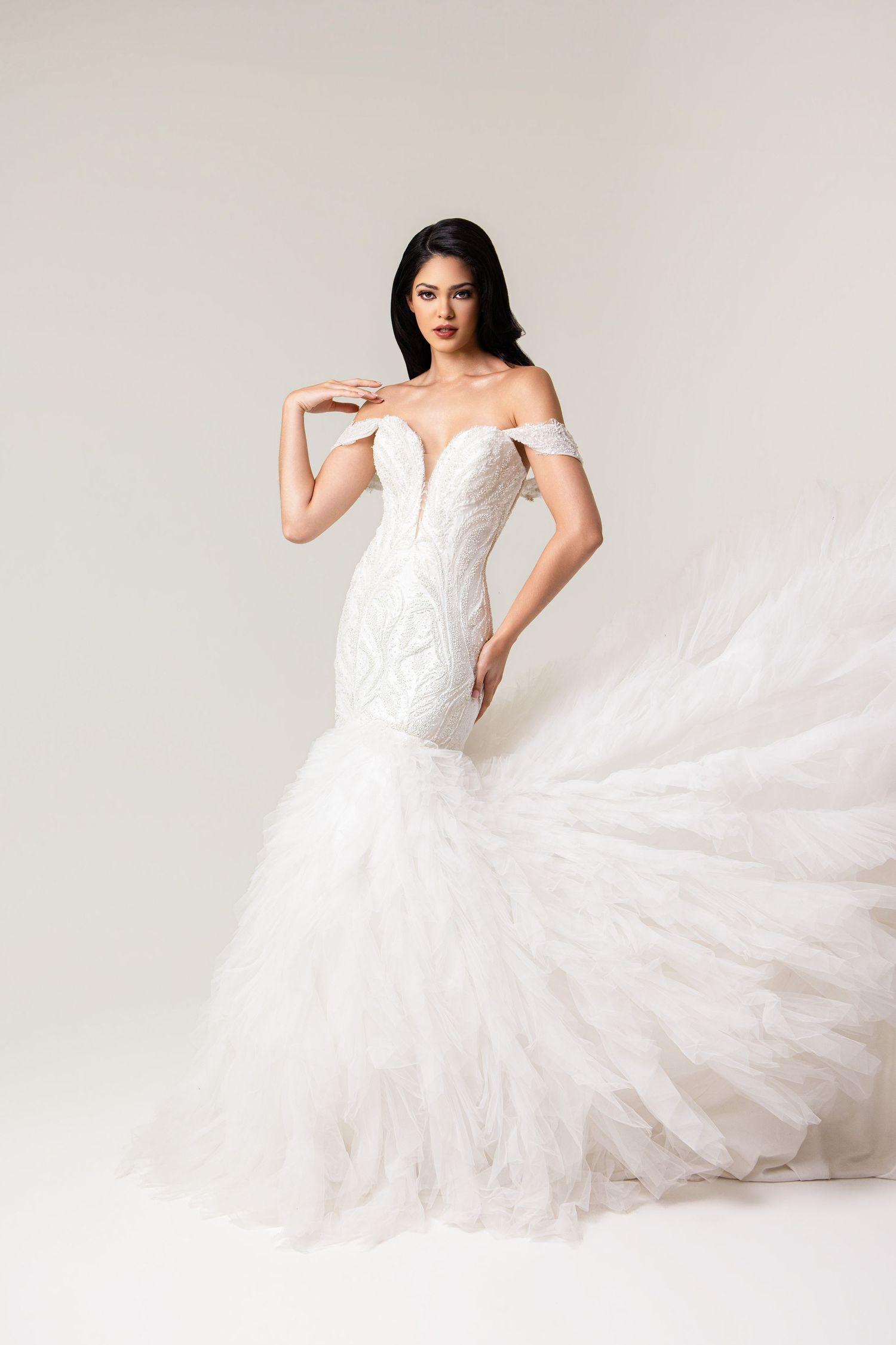 Woman in elegant off-shoulder mermaid wedding gown with lace bodice and flowing tulle skirt on white background.