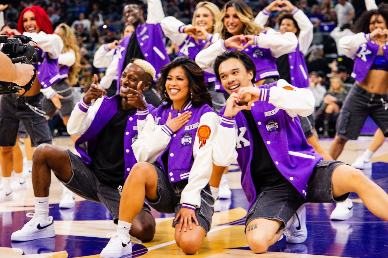 NBA cheerleaders in purple varsity jackets pose on court with fans during a Sacramento Kings game.