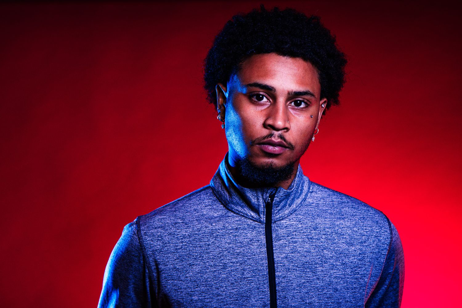 Young Black man with afro hair wearing a blue athletic jacket poses against a bold red background.