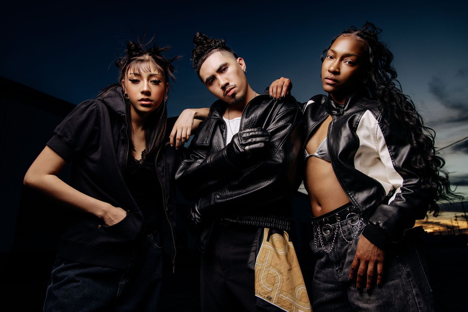 Fashion models pose dramatically in dark lighting wearing black leather and denim outfits for an edgy editorial shoot.