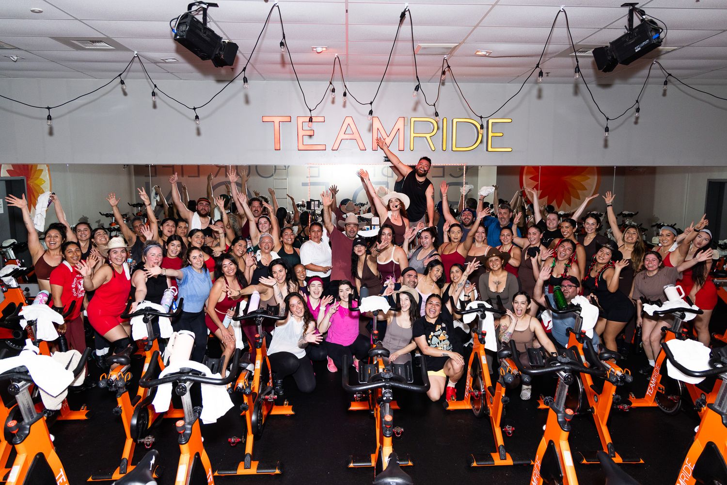Large group of dancers perform synchronized dance moves in a dimly lit room with string lights overhead.