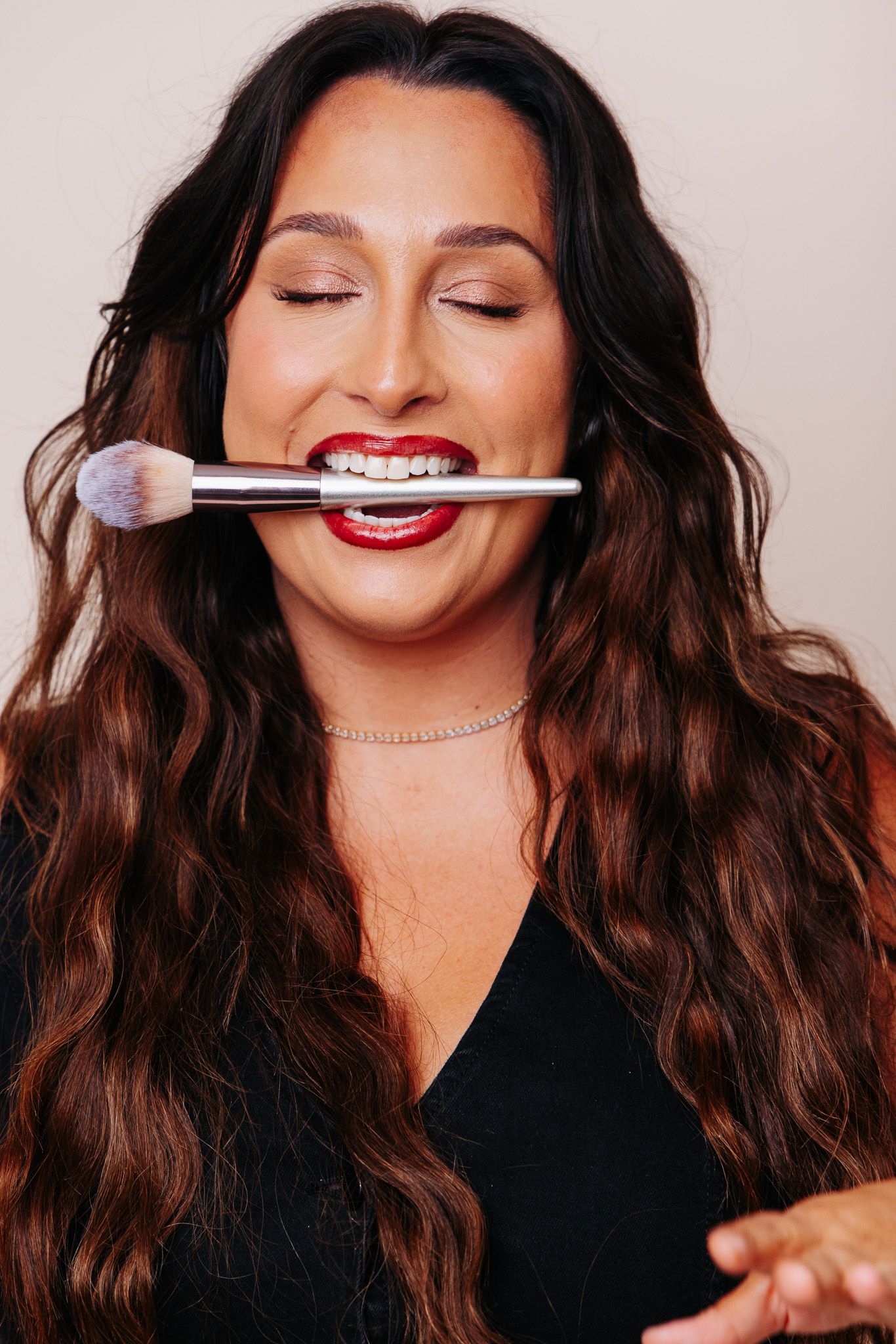 Woman with long wavy hair holds makeup brush in her teeth, wearing red lipstick and black top.
