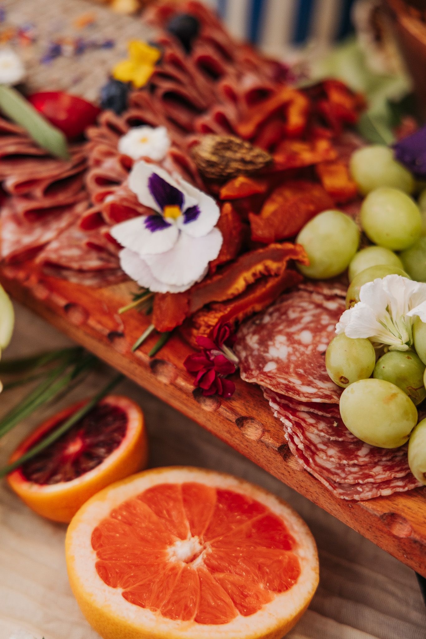 An elegant charcuterie board featuring sliced meats, fresh grapefruits, green grapes, and edible flowers on a wooden surface.
