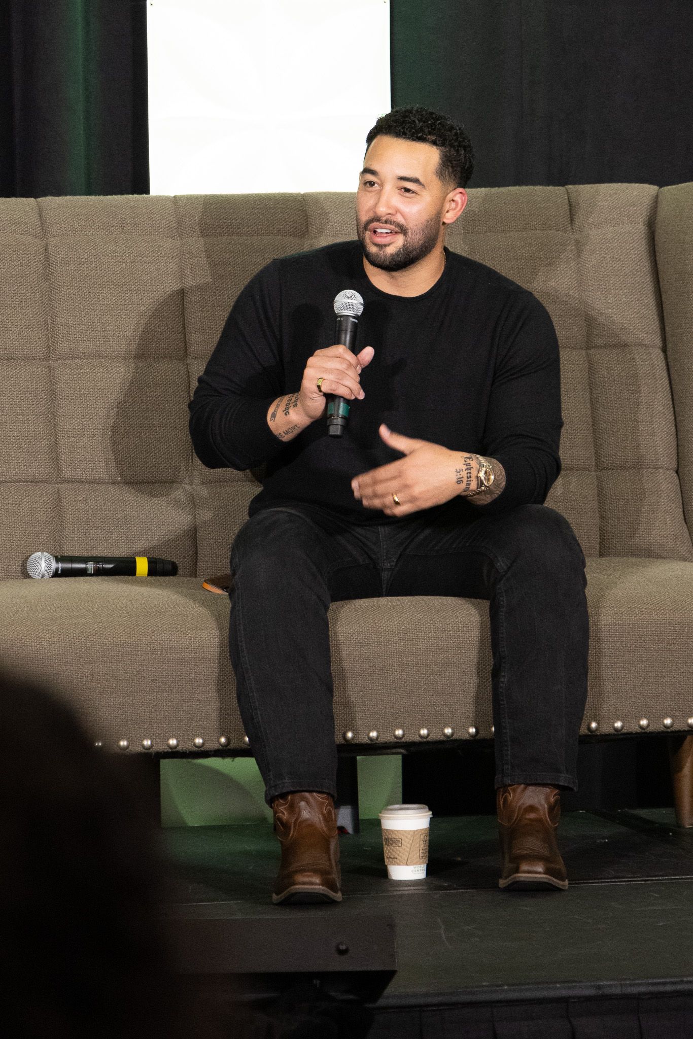 A man wearing a black shirt sits on a gray couch speaking into a microphone during what appears to be a panel discussion.