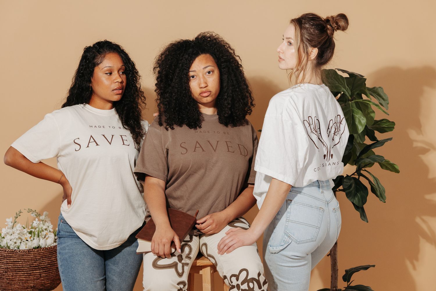 Two women in casual t-shirts and denim posing against a beige wall in a natural light studio setting.