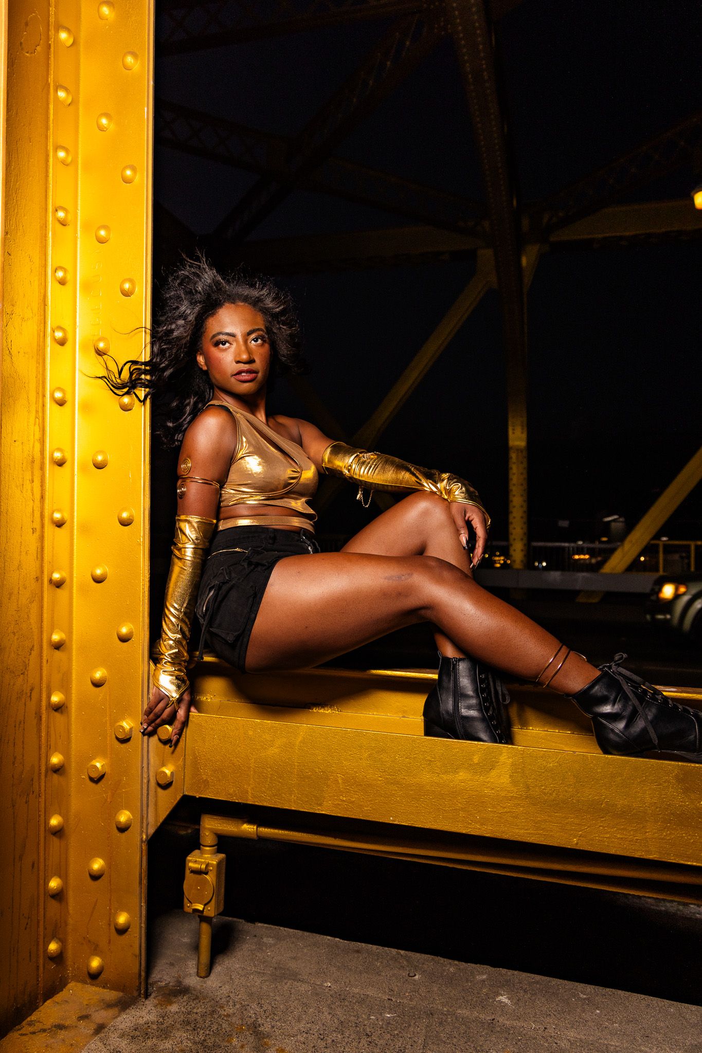 Fashion model poses dramatically on yellow industrial beam in dark moody setting.