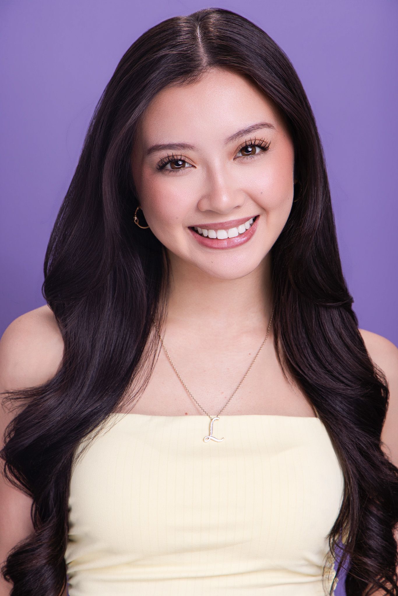 Young woman with long dark hair smiling, wearing yellow top and initial necklace against purple background.